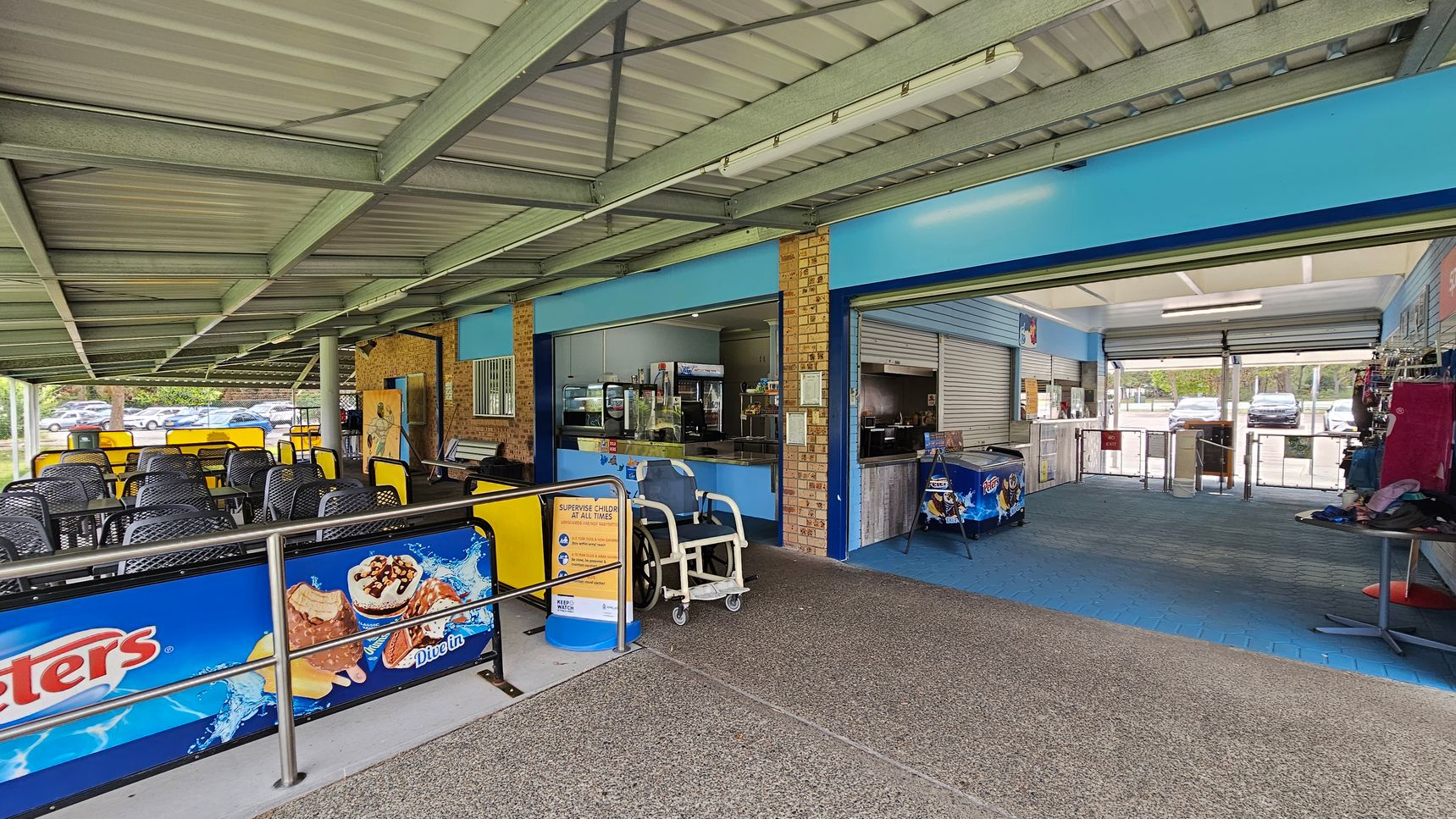A covered outdoor seating area with black and yellow chairs on the left. The floor is a mix of textured concrete and blue tiles. In the centre, there's a counter with a display of snacks and drinks. On the right, an open area with a blue-tiled floor leads to a glass entrance with turnstiles. Ice cream advertisements are visible on barriers and a freezer. The ceiling is metal with exposed beams. A covered outdoor seating area with black and yellow chairs on the left. The floor is a mix of textured concrete and blue tiles. In the centre, there's a counter with a display of snacks and drinks. On the right, an open area with a blue-tiled floor leads to a glass entrance with turnstiles. Ice cream advertisements are visible on barriers and a freezer. The ceiling is metal with exposed beams.