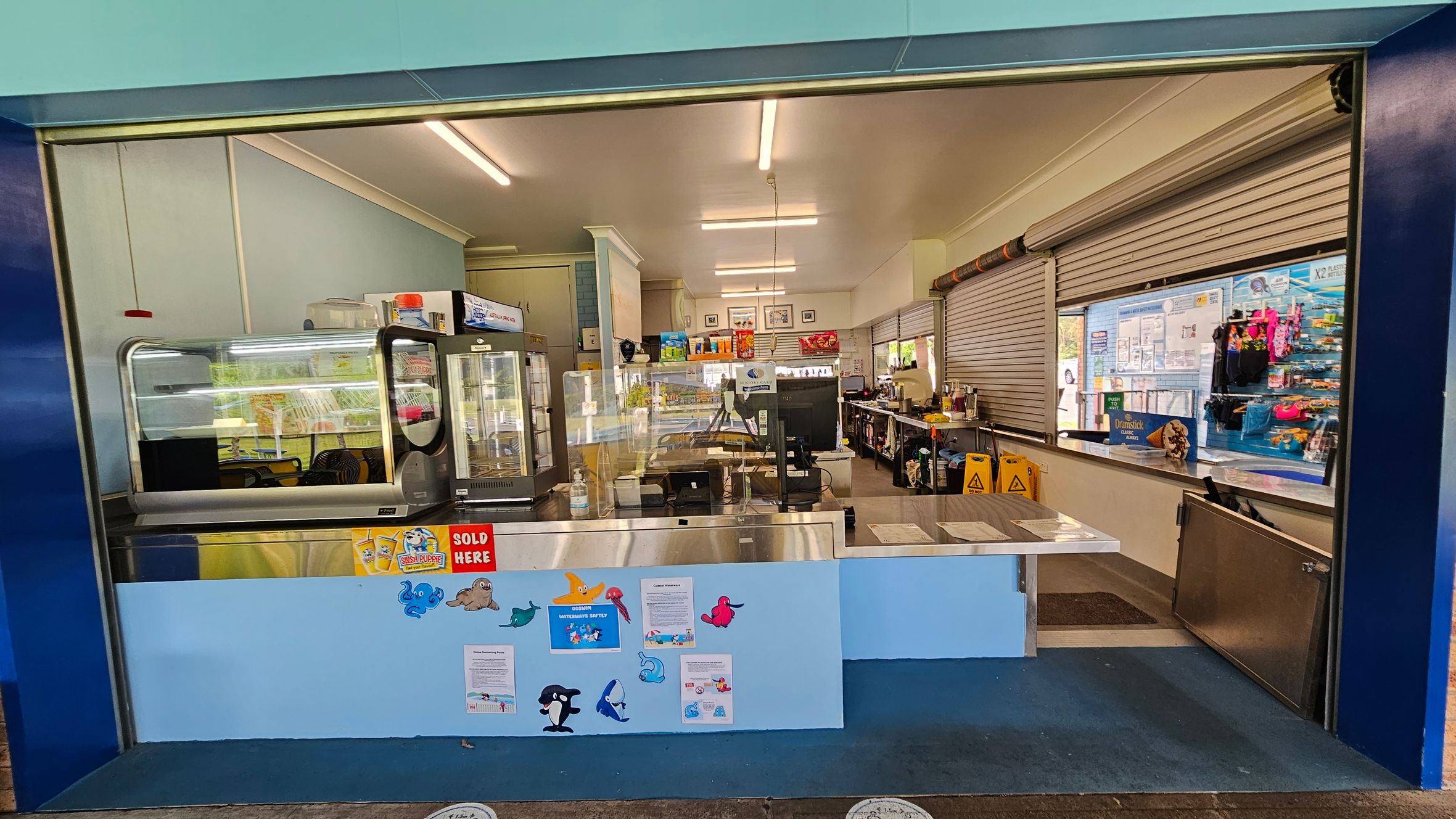 A canteen with a wide serving counter in the foreground, decorated with colourful sea creature cutouts and signs. Behind the counter, there are display fridges and shelves stocked with various items. The interior is well-lit with fluorescent lights, and the walls are light blue. On the right, there are closed roller shutters and a display of snacks and drinks. The floor is blue, and there are caution signs visible inside. A canteen with a wide serving counter in the foreground, decorated with colourful sea creature cutouts and signs. Behind the counter, there are display fridges and shelves stocked with various items. The interior is well-lit with fluorescent lights, and the walls are light blue. On the right, there are closed roller shutters and a display of snacks and drinks. The floor is blue, and there are caution signs visible inside.