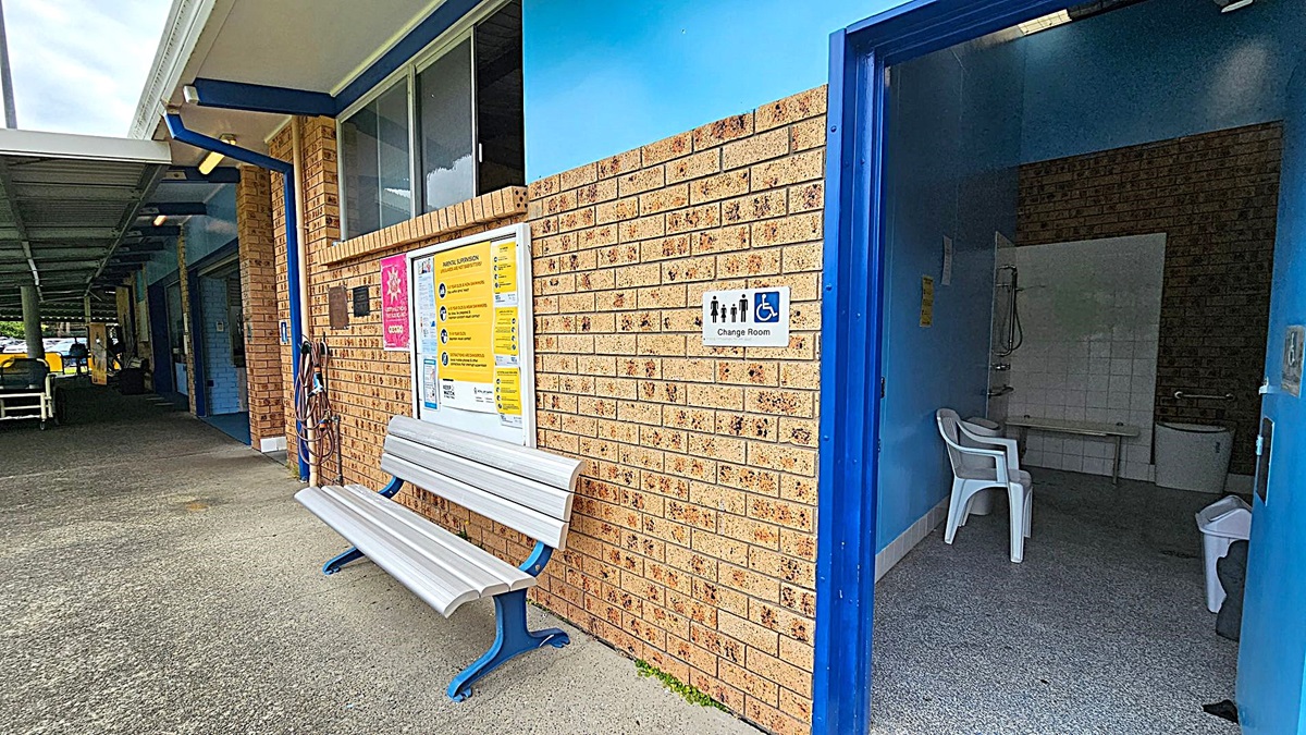 A brick building with a blue trim features a change room on the right, marked by a sign with accessibility symbols. Inside, there's a white plastic chair and a tiled area with a shower. On the left, a white bench with blue legs is positioned against the wall, which displays various informational posters. The floor is concrete, and a covered walkway extends into the background. A brick building with a blue trim features a change room on the right, marked by a sign with accessibility symbols. Inside, there's a white plastic chair and a tiled area with a shower. On the left, a white bench with blue legs is positioned against the wall, which displays various informational posters. The floor is concrete, and a covered walkway extends into the background.
