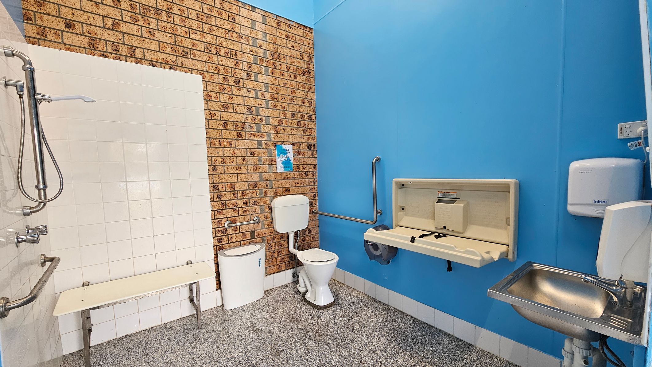 A bathroom with a brick wall on the left and a blue wall on the right. The left side features a shower with a metal handle and a white tiled wall, along with a foldable bench. In the centre, there's a toilet with a white cistern and a small bin beside it. The right side has a baby changing station, a stainless steel sink, and a wall-mounted hand dryer. The floor is speckled grey, and there are grab bars near the toilet and changing station. A bathroom with a brick wall on the left and a blue wall on the right. The left side features a shower with a metal handle and a white tiled wall, along with a foldable bench. In the centre, there's a toilet with a white cistern and a small bin beside it. The right side has a baby changing station, a stainless steel sink, and a wall-mounted hand dryer. The floor is speckled grey, and there are grab bars near the toilet and changing station.