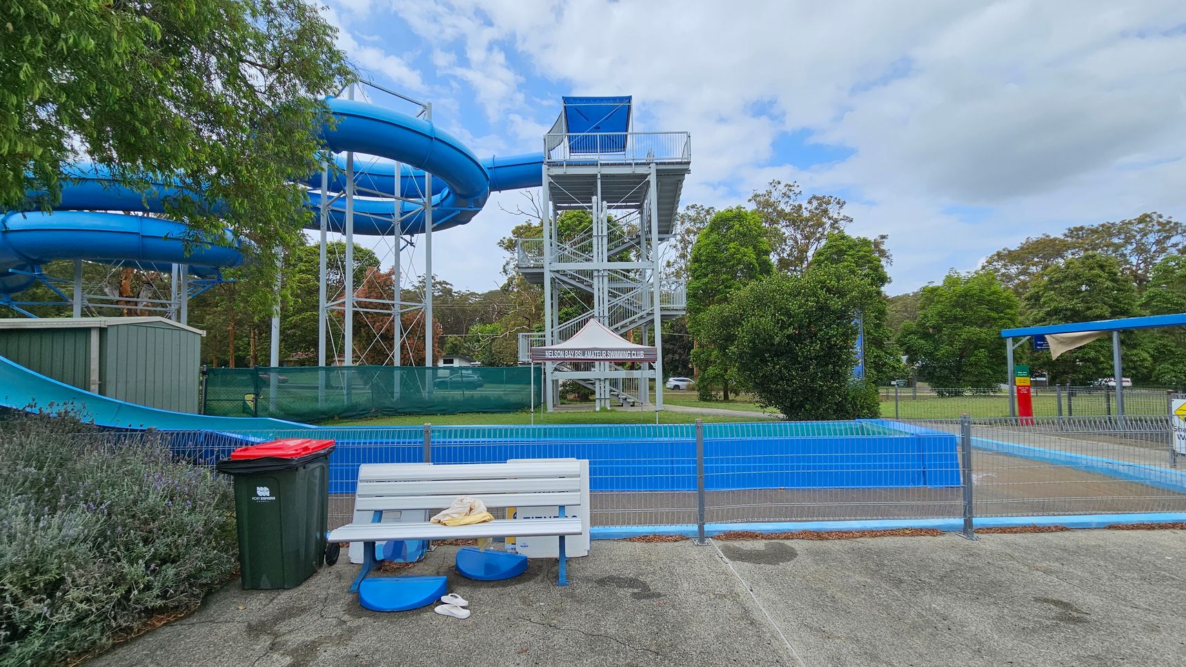 A water park scene with a large blue water slide on the left, supported by a metal structure. In the centre, there's a tall platform with stairs leading up to the slide. A sign reads "Nelson Bay Swimming Club" under a small canopy. In the foreground, a white bench with a towel and flip-flops is next to a green bin with a red lid. The area is fenced, and there are trees and grass in the background. A small shelter with a shade cloth is on the right. A water park scene with a large blue water slide on the left, supported by a metal structure. In the centre, there's a tall platform with stairs leading up to the slide. A sign reads "Nelson Bay Swimming Club" under a small canopy. In the foreground, a white bench with a towel and flip-flops is next to a green bin with a red lid. The area is fenced, and there are trees and grass in the background. A small shelter with a shade cloth is on the right.