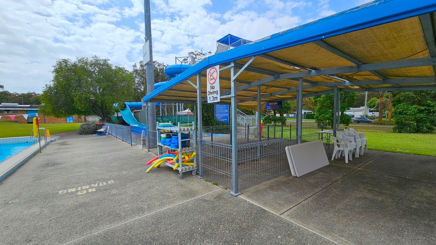 A poolside area with a blue water slide on the left, surrounded by a fence. A paved walkway leads to a covered seating area on the right, with stacked white plastic chairs and a folded table. A sign reads "No Diving 1.3m". Pool equipment, including colourful noodles, is stored on a rack. In the background, there are trees and a grassy area. The ceiling of the covered area is made of a beige material supported by a metal frame. A poolside area with a blue water slide on the left, surrounded by a fence. A paved walkway leads to a covered seating area on the right, with stacked white plastic chairs and a folded table. A sign reads "No Diving 1.3m". Pool equipment, including colourful noodles, is stored on a rack. In the background, there are trees and a grassy area. The ceiling of the covered area is made of a beige material supported by a metal frame.