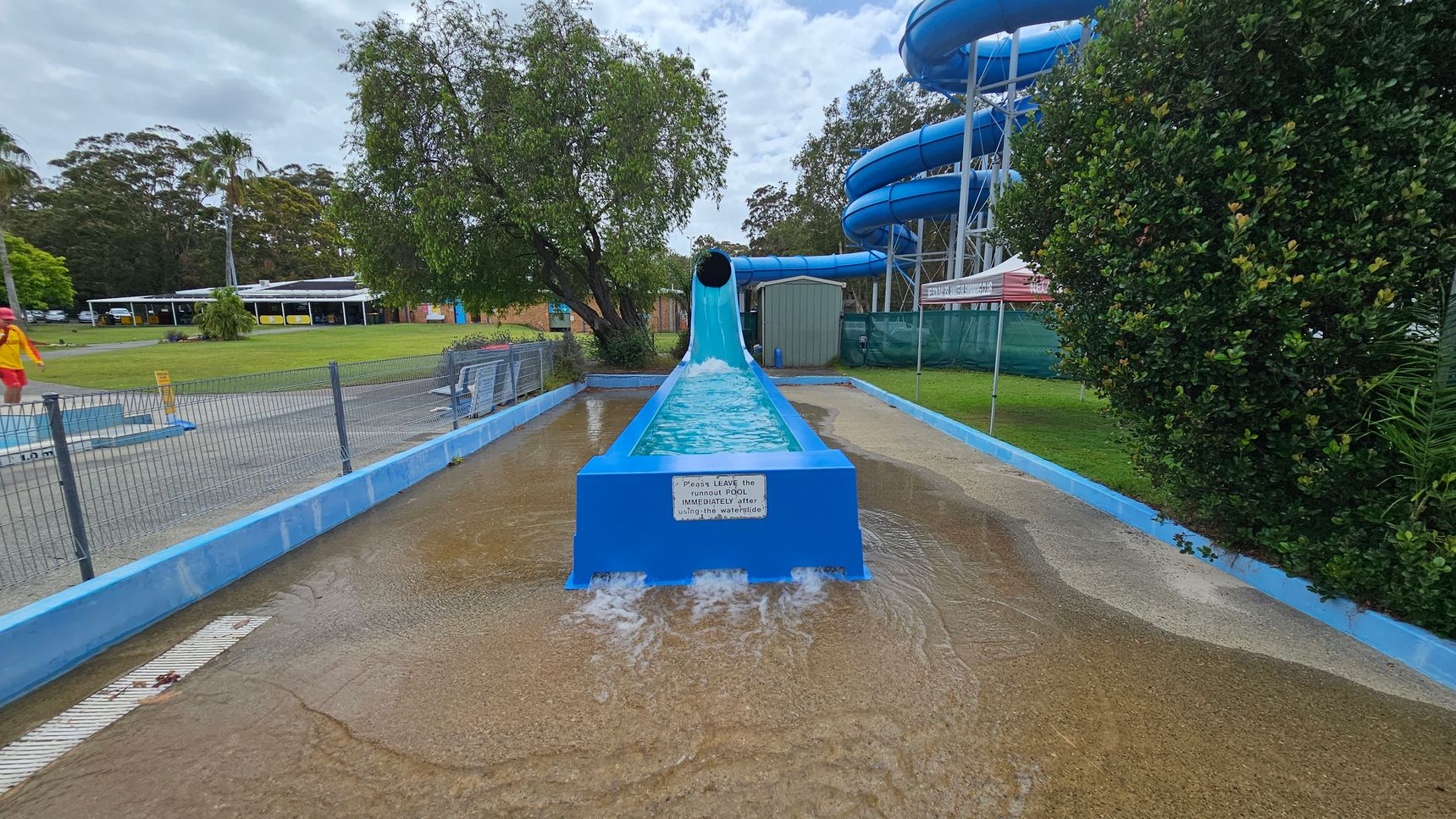 A blue water slide with a tunnel at the top ends in a shallow pool. Water flows out of the slide into the pool. On the left, a lifeguard in a red and yellow uniform walks near a fenced area. In the background, there are trees and a building with a white roof. On the right, a large bush and a blue spiral slide structure are visible. The ground is wet, and there is a sign on the slide's end. A blue water slide with a tunnel at the top ends in a shallow pool. Water flows out of the slide into the pool. On the left, a lifeguard in a red and yellow uniform walks near a fenced area. In the background, there are trees and a building with a white roof. On the right, a large bush and a blue spiral slide structure are visible. The ground is wet, and there is a sign on the slide's end.
