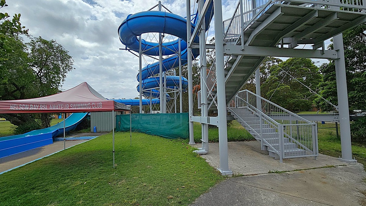 On the left, a blue water slide spirals down from a tall metal structure. Below it, a tent with "Nelson Bay RSL Amateur Swimming Club" written on it is set up on a grassy area. On the right, a metal staircase leads up to the slide, with a concrete path at its base. Trees and greenery are visible in the background, providing a natural setting around the water slide and tent. On the left, a blue water slide spirals down from a tall metal structure. Below it, a tent with "Nelson Bay RSL Amateur Swimming Club" written on it is set up on a grassy area. On the right, a metal staircase leads up to the slide, with a concrete path at its base. Trees and greenery are visible in the background, providing a natural setting around the water slide and tent.