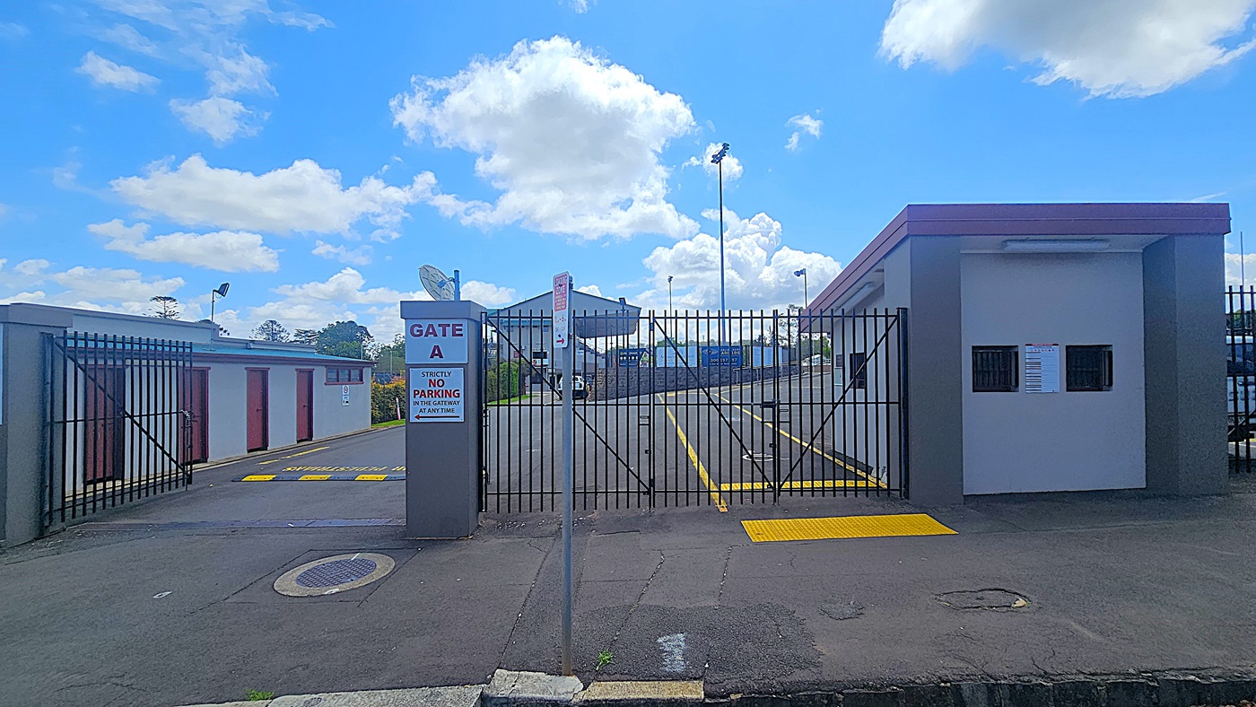 A gated entrance labelled "Gate A" with a sign stating "Strictly No Parking in the Gateway at Any Time" is in the centre. The gate is flanked by a small building on the right with two small windows and a notice. On the left, there are several storage units with brown doors. The ground is paved with yellow markings, and a street sign is visible. In the background, there are tall floodlights and a partially visible structure. The sky is blue with scattered clouds.