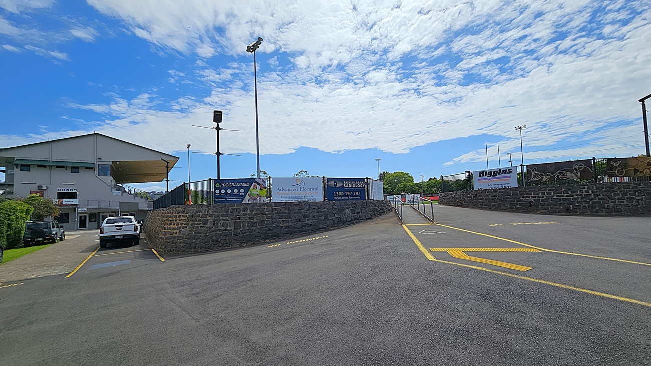 A paved area with yellow markings leads to a sports facility. On the left, a white vehicle is parked near a building with a sloped roof and several signs. In the centre, a stone wall with advertisements lines the path. On the right, a fenced area with more signs and a glimpse of a sports field is visible. Tall floodlights are positioned throughout the area.