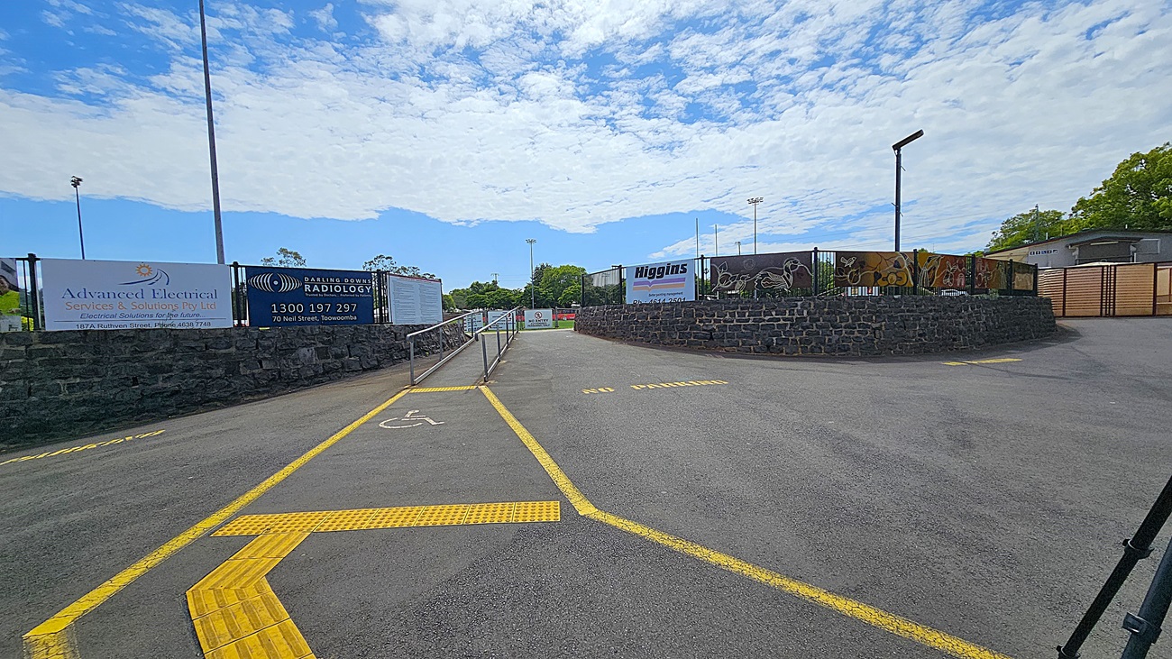 A paved area with a ramp in the centre, marked with yellow lines and a wheelchair symbol. On the left, there are signs for "Advanced Electrical" and "Darling Downs Radiology" attached to a stone wall. On the right, a curved stone wall with decorative fencing and a sign for "Higgins". Trees and a building are visible in the background. A tall light pole and a streetlamp are also present.