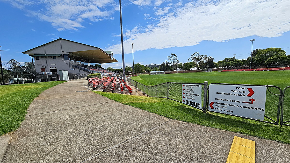 A sports stadium with a covered grandstand on the left, featuring rows of red seats. A concrete path runs alongside the stand, bordered by green grass. On the right, a large grassy field is visible, with a sign in the foreground providing directions to various stands, food, and toilets. Trees and additional seating are in the background. The sky is partly cloudy.