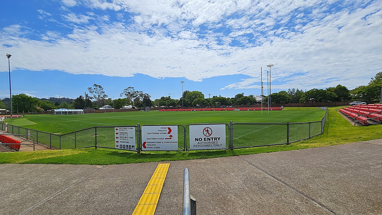 A sports field with a green grass pitch is surrounded by a chain-link fence. In the foreground, a concrete path leads to a gate with signs indicating "No Entry" and directions to various stands and facilities. On the right, there are rows of red stadium seats. Trees and a few buildings are visible in the background, along with tall floodlights. The sky is partly cloudy.