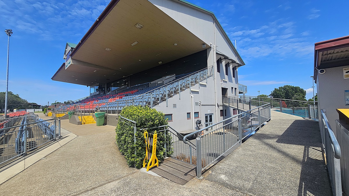 On the left, there is a large grandstand with rows of red and blue seats, partially covered by a sloping roof. A concrete pathway runs alongside, bordered by metal railings and a green hedge. On the right, a wide ramp with metal railings leads upwards, flanked by a small building with a red roof. The foreground features a concrete floor, while the ceiling of the grandstand is visible, with several lights attached.