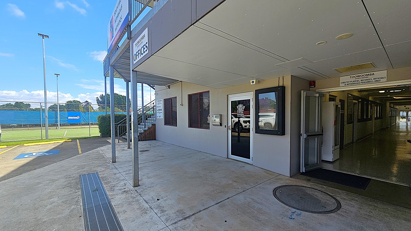 On the left, there is a sports field with a blue fence and tall floodlights. A concrete path with a disabled parking space leads to the building. On the right, a corridor extends into the distance, lined with windows and an open door. In the middle, a building entrance with glass doors and a sign above reads "Toowoomba Offices". The floor is concrete with a metal drainage cover, and the ceiling is flat with recessed lighting.