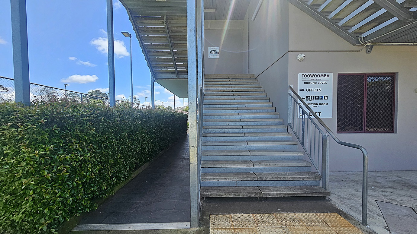 On the left, a pathway lined with dense green hedges runs parallel to a metal fence with tall floodlights. On the right, a wide staircase with metal railings leads up to a building entrance. A sign on the wall indicates directions to various offices. The foreground features a textured concrete floor with tactile paving near the stairs. The ceiling above the stairs is flat with a fluorescent light fixture.