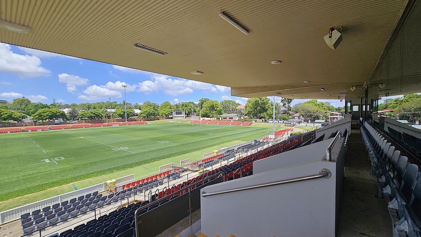 On the left, a green sports field with white markings and red seating along the perimeter. On the right, rows of dark blue seats under a covered grandstand. In the middle, a walkway with metal railings leads downwards. The background features trees and small buildings. The floor is concrete, and the ceiling of the grandstand is beige with recessed lighting and speakers.