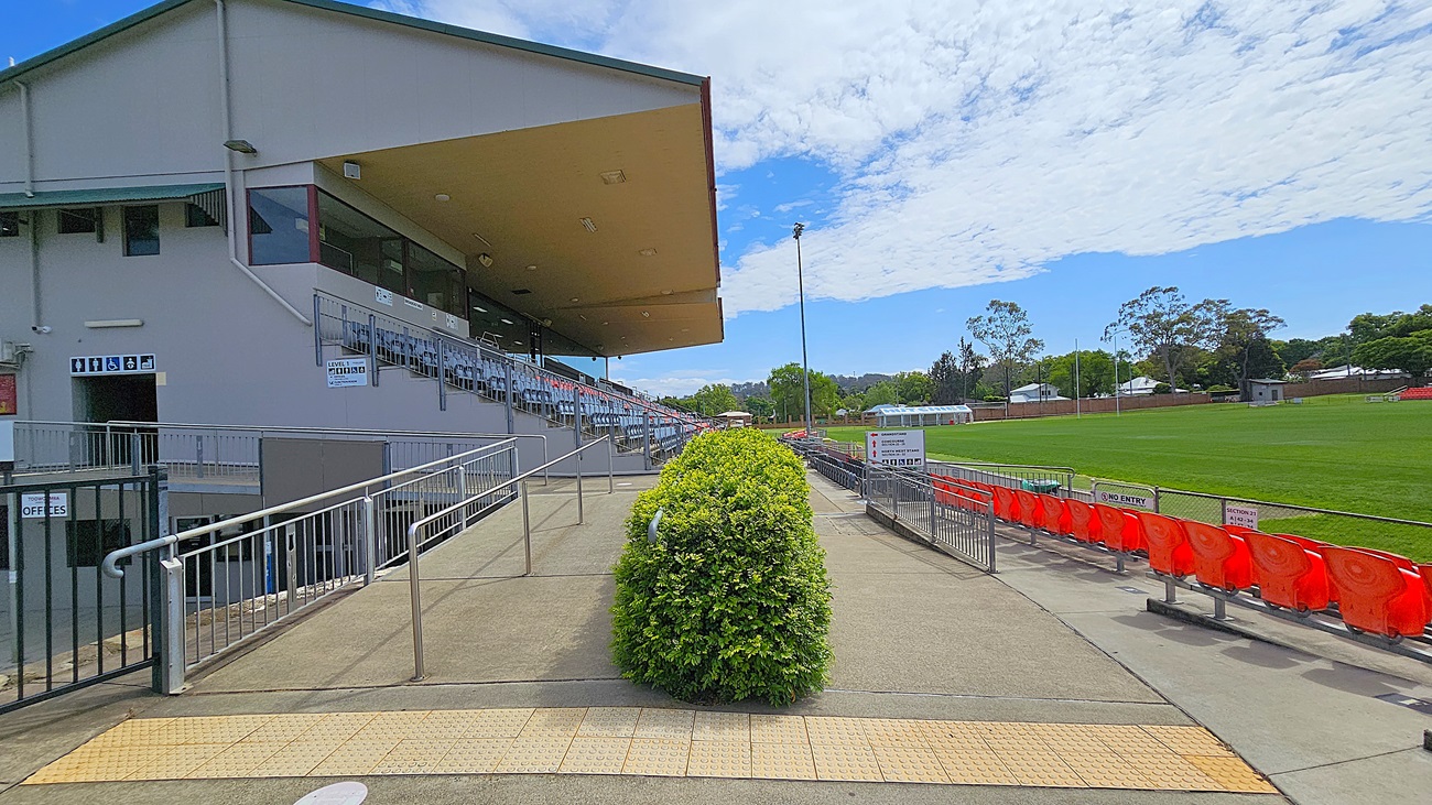 A sports stadium with a covered seating area on the left, featuring rows of blue seats. A ramp with metal railings leads up to the seating area. In the centre, a neatly trimmed green bush lines the walkway. On the right, there are red plastic seats facing a green sports field. Signs indicate "No Entry" and "Offices". The ground is paved, and the ceiling of the covered area is visible. Trees and houses are in the background.