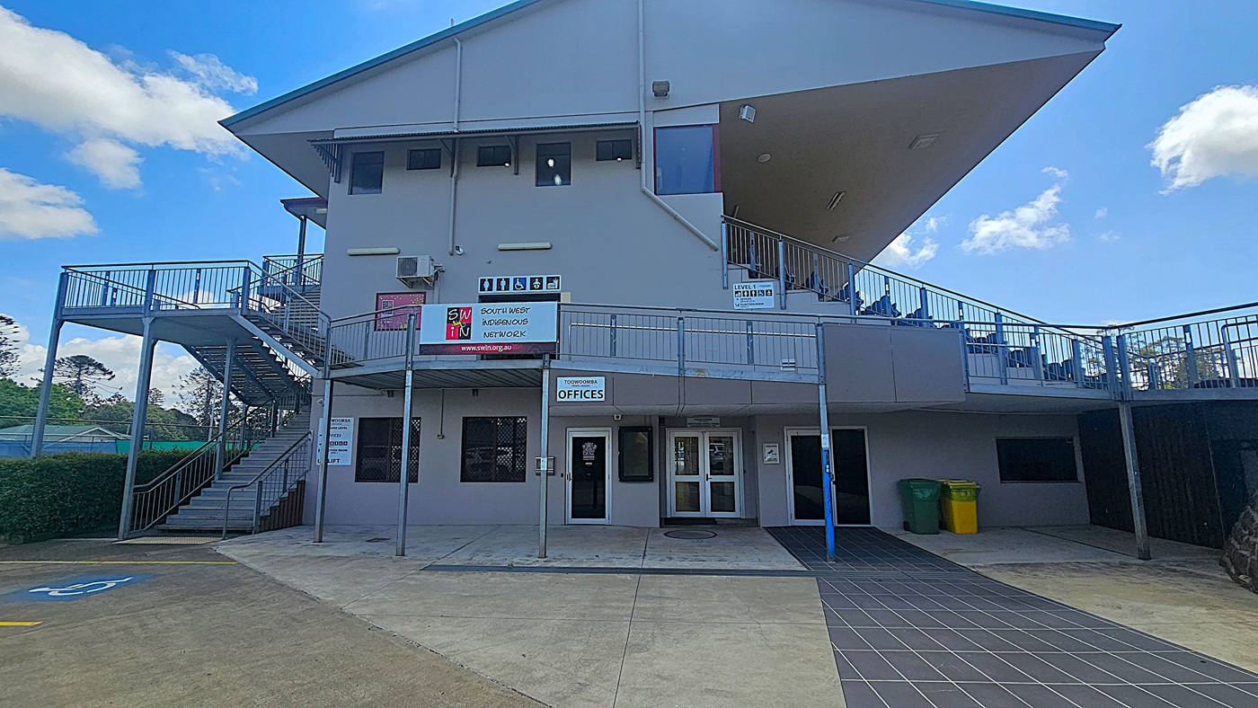 A two-storey building with a sloped roof. On the left, there is an external staircase leading to the upper level. The middle section has signs for the South West Indigenous Network and Toowoomba Offices. The ground level has several doors and windows. On the right, there are two bins, one green and one yellow. The foreground features a paved walkway and a parking area with a marked accessible parking space. Trees are visible in the background.
