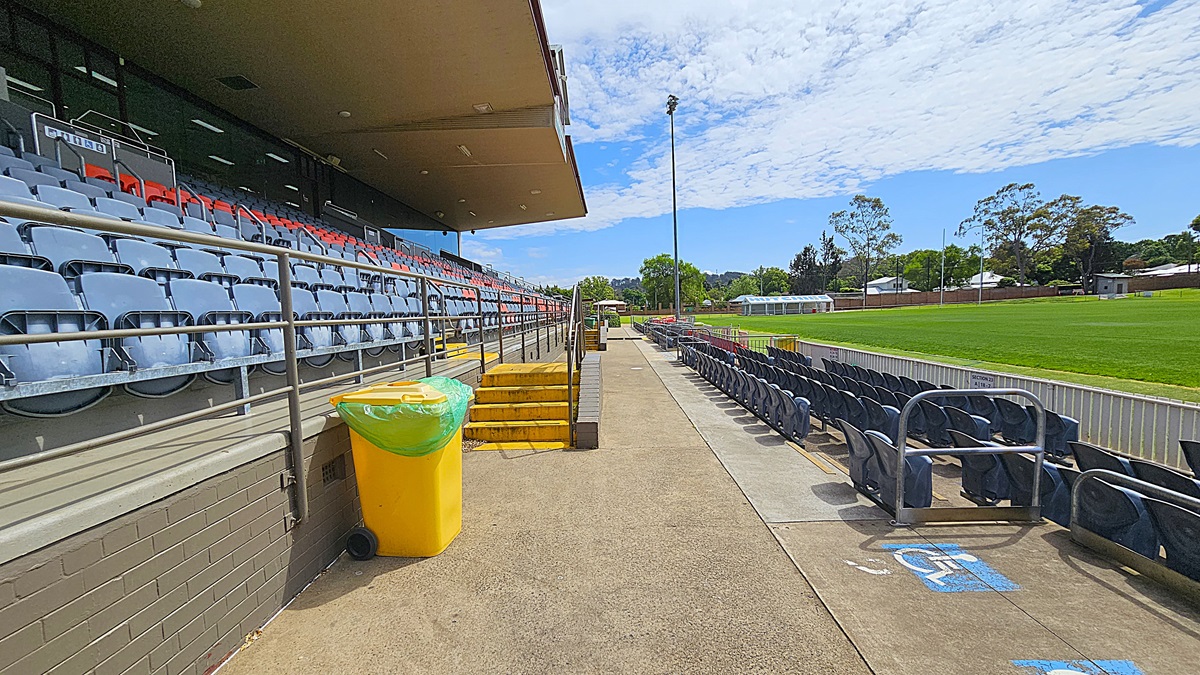 On the left, there are rows of blue and red stadium seats under a covered area, with a yellow bin nearby. A set of yellow steps leads down to a walkway. On the right, more seating is visible, with a section marked for accessibility. In the background, a green sports field is surrounded by trees and a few buildings. A tall floodlight stands near the field. The ground is paved, and the ceiling of the covered area is flat and extends outward.