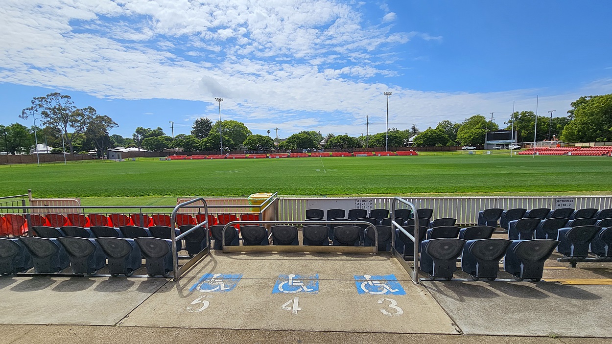 A sports field with a well-maintained green pitch is surrounded by red seating. In the foreground, there are black seats and a section marked for wheelchair access with blue symbols on the concrete. Trees and a few buildings are visible in the background, along with tall floodlights. A scoreboard is on the right side. The sky is partly cloudy.
