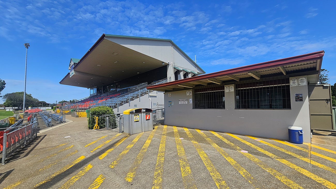 A sports stadium with a large covered grandstand on the left, featuring red and blue seating. In the foreground, a concrete path with yellow painted lines leads towards the grandstand. On the right, a small building with a red roof and barred windows is visible, with signs and a blue bin nearby. Metal railings line the path, and a tall floodlight stands in the background. Trees and a grassy area are visible to the left.