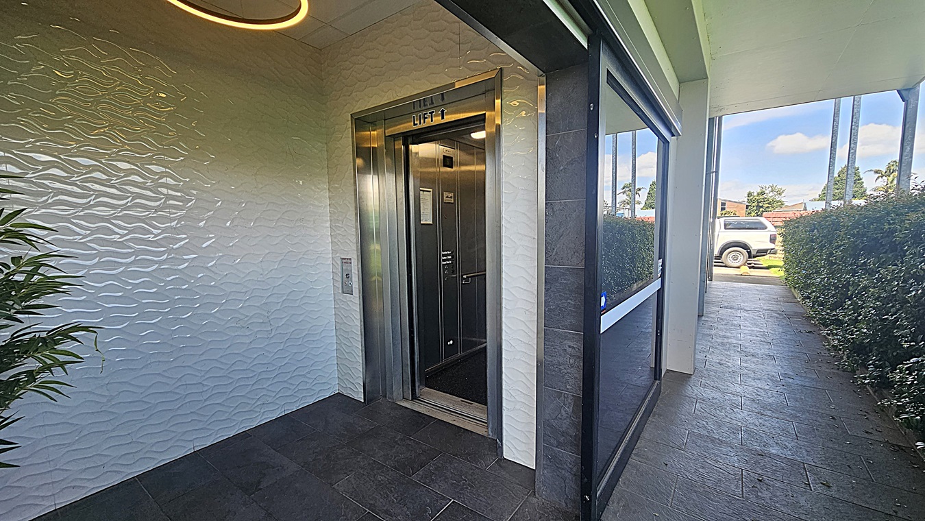 A lift entrance is on the left, with metallic doors and a sign above reading "LIFT". The wall has a textured wave pattern. A plant is visible in the corner. On the right, there's a glass window reflecting the outside. The floor is tiled in dark grey. In the background, a pathway leads to an outdoor area with a white vehicle parked and green hedges lining the path.