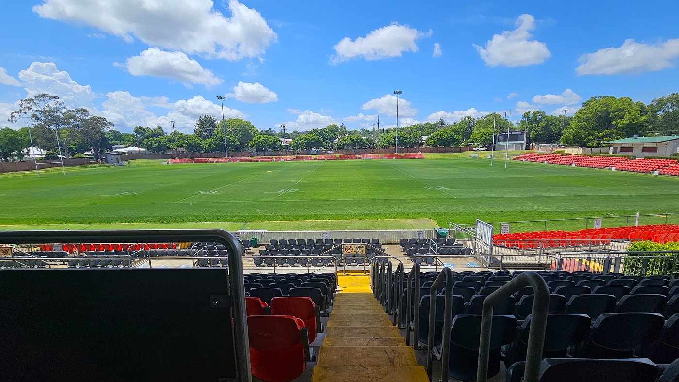 View from the stands of a rugby field. In the foreground, rows of black and red seats are visible, with a central aisle leading down to the field. The field is lush and green, marked with white lines. Surrounding the field are more red seats and goalposts at each end. In the background, there are trees and some buildings, with a clear blue sky above.