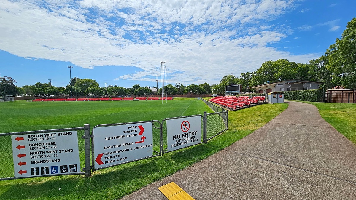 A sports field with green grass is on the left, surrounded by a fence with signs indicating directions to various stands and facilities. Red seating is visible in the background. On the right, a concrete path leads towards a building partially obscured by trees and hedges. A "No Entry" sign is attached to the fence. The sky is partly cloudy.