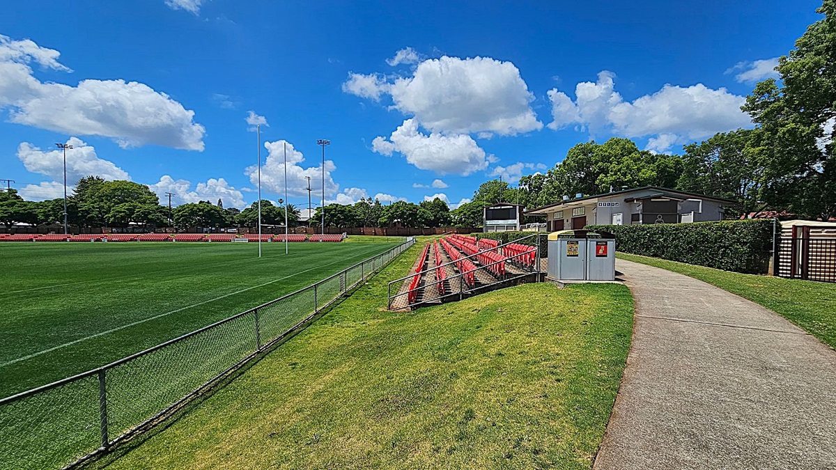 A sports field with green grass and a metal fence on the left. Red seating is arranged in rows on a grassy slope to the right. A concrete path runs alongside the seating, leading to a building partially obscured by trees and hedges. A utility box is near the path. Floodlights and goalposts are visible on the field.