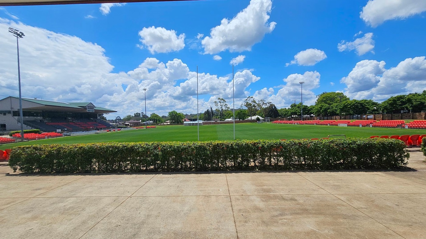 A sports field with a well-maintained green pitch is in the centre, surrounded by red seating. On the left, there is a large grandstand with a roof and more seating. Tall floodlights are visible on both sides. In the foreground, a hedge runs along the edge of a concrete walkway. Trees and buildings are in the background, providing a natural border around the stadium.