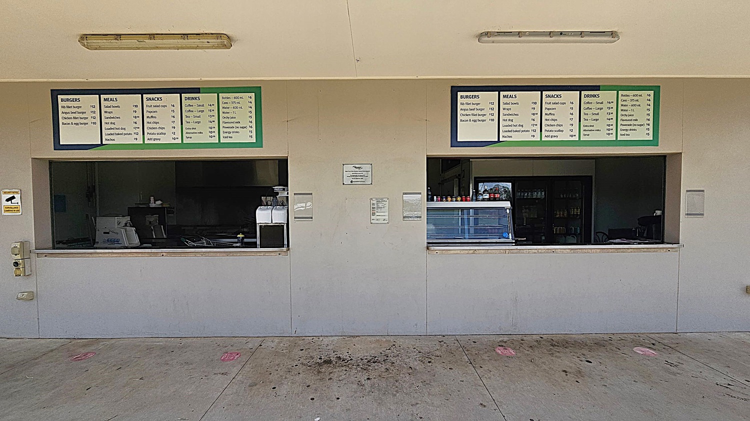A canteen with two service windows. Above each window are menu boards listing items like burgers, meals, snacks, and drinks. The left window is closed, with a coffee machine visible inside. The right window has a display case and condiment bottles. The wall is light-coloured, and the floor is concrete with some stains. Two fluorescent lights are mounted on the ceiling.