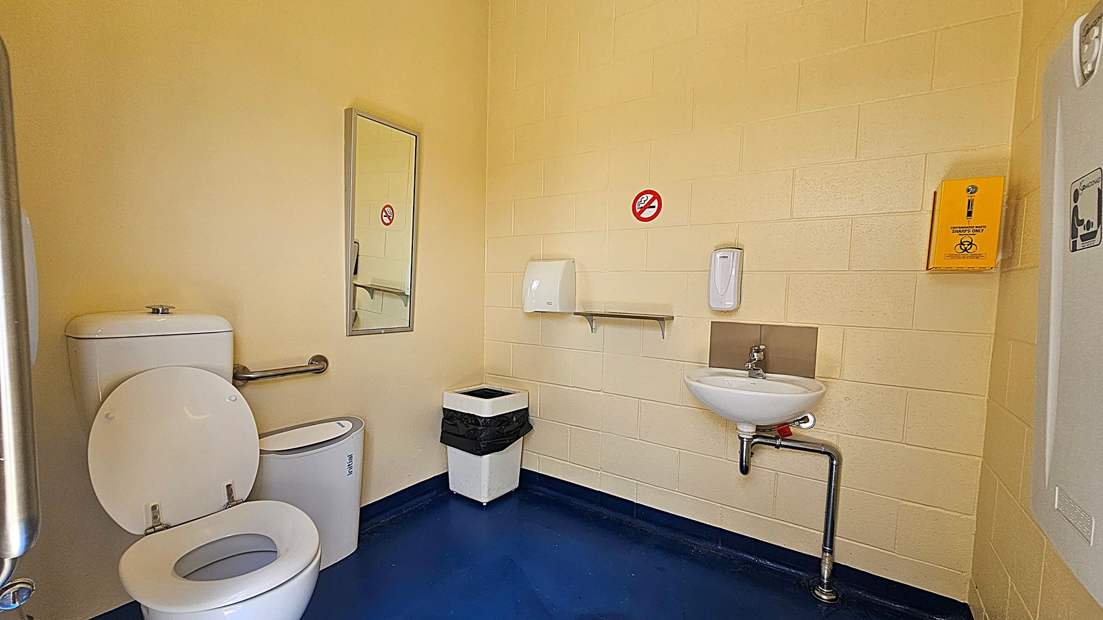 A public toilet with a white toilet and a grab rail on the left. A mirror is mounted on the wall above the toilet. In the centre, there's a bin with a black liner. On the right, a small white sink with a soap dispenser above it. A paper towel dispenser and a no smoking sign are on the wall. A yellow sharps disposal box is mounted on the wall. The floor is dark blue, and the walls are light yellow.