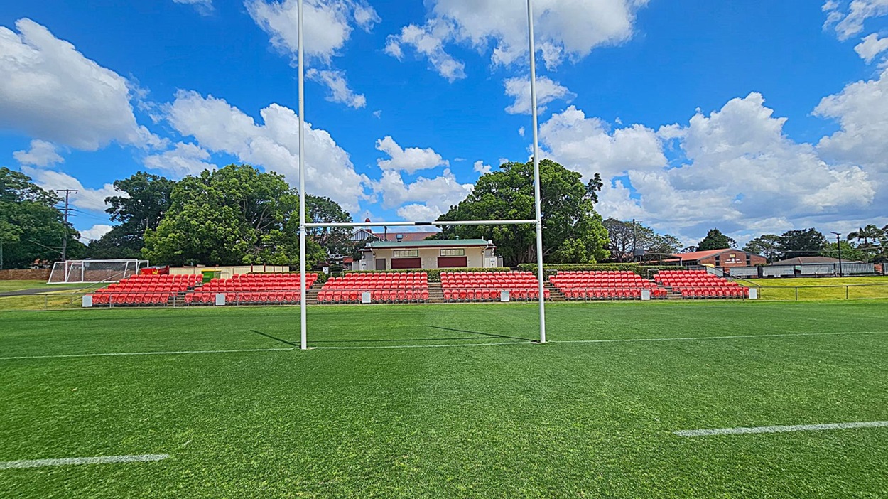 A rugby field with green grass and white goalposts in the foreground. In the background, there are rows of red stadium seats. Behind the seats, there are trees and a building with a beige facade and a red roof. To the left, a football goalpost is visible. The sky is blue with scattered white clouds.