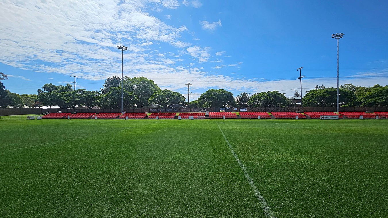 A football field with lush green grass in the foreground. In the background, there are rows of red seats, partially shaded by trees. Tall floodlights are positioned on either side of the seating area. The trees are dense and provide a natural backdrop to the scene.