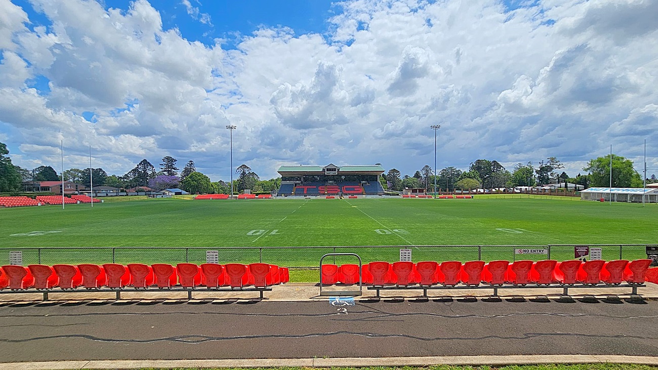 A rugby field with green grass and white markings. In the foreground, there are rows of red plastic seats. The background features a grandstand with a roof and more seating, displaying the letters "TSC" in red. Tall floodlights are positioned around the field, and there are goalposts at each end. Trees and buildings are visible beyond the field.