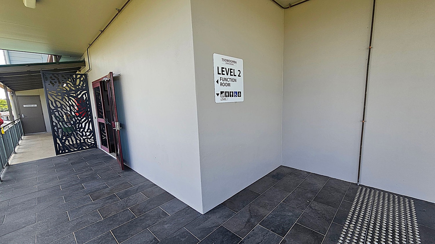 A corridor with dark tiled flooring and a light-coloured wall. On the right, a sign reads "Toowoomba Level 2 Function Room" with directional arrows and icons. To the left, there's a decorative metal screen and a door with glass panels. Further left, a grey door marked "Camera Room" is visible. The ceiling is plain with visible piping.