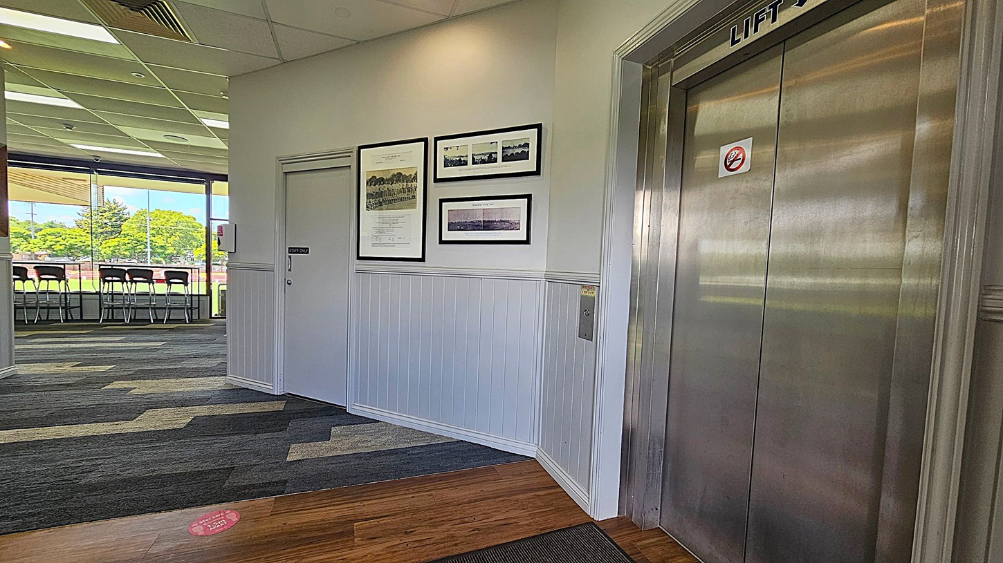 A hallway with a lift on the right, featuring metallic doors and a "Lift" sign above. On the left, a "Staff Only" door is surrounded by framed photographs and documents on the wall. The floor is a mix of wood and patterned carpet. In the background, large windows reveal a view of green trees and a sports field with chairs lined up against the glass. The ceiling has recessed lighting and air vents.