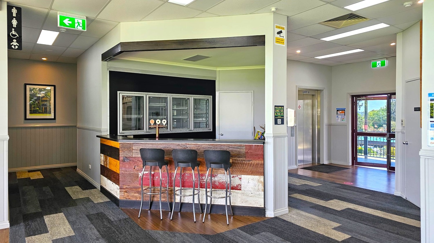 A modern interior with a small bar area in the centre, featuring a rustic wooden counter and three high stools. Behind the bar are glass-fronted cabinets. To the left, a corridor leads to a framed picture on the wall and a sign indicating accessible toilets. On the right, there's a lift and a glass door leading outside, showing a view of greenery. The floor is covered with patterned carpet tiles, and the ceiling has recessed lighting and exit signs.
