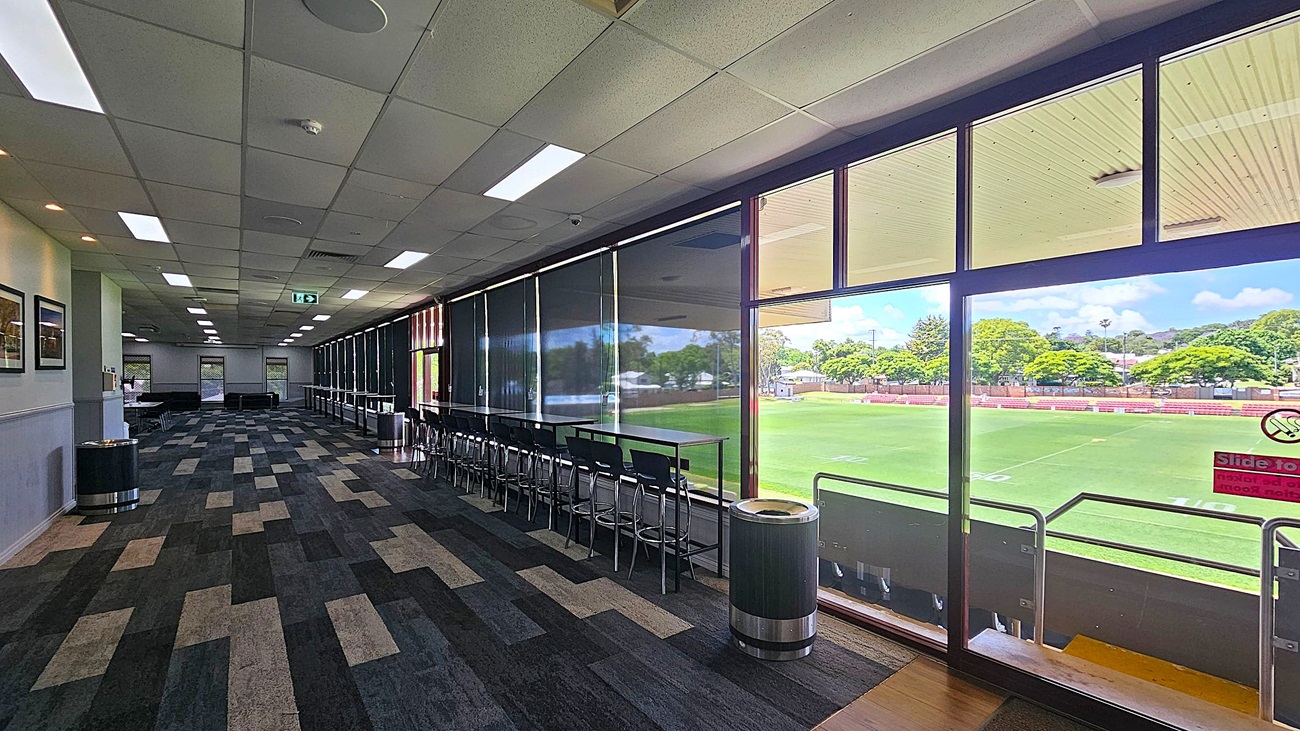 A spacious room with a patterned carpet floor and a ceiling with recessed lighting. On the left, there are framed pictures on the wall and a waste bin. The right side features large windows with blinds, offering a view of a green sports field and trees outside. In front of the windows, there are high tables and stools. The room extends into the distance with more seating and tables visible.