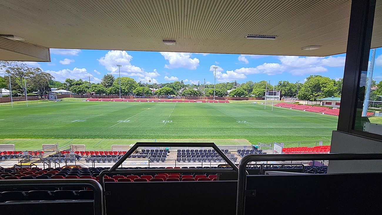 View from a covered stadium seating area overlooking a rugby field. The foreground has rows of red and blue seats. The field is marked with white lines and goalposts at each end. In the background, there are red bleachers and a scoreboard. Trees and a few buildings are visible beyond the field. The ceiling above the seating area is lined with panels and lights.
