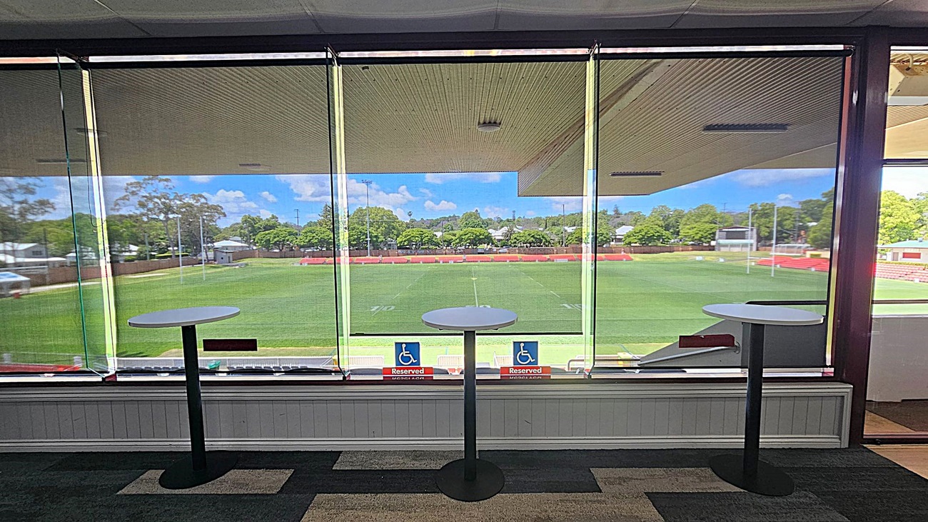 A view from inside a building with large glass windows overlooking a green sports field. Three tall round tables with black bases are positioned in front of the windows. The floor is carpeted in a dark pattern. Outside, there are trees and a few small structures in the background. Two signs on the window indicate reserved seating for accessibility. The ceiling inside is light-coloured with a textured pattern.