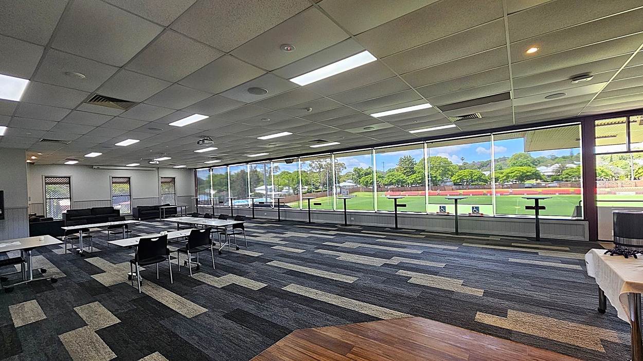 A spacious room with a patterned carpet in shades of grey and green. On the left, there are several tables and chairs arranged in a meeting setup. The ceiling is fitted with fluorescent lights and air vents. The right side features large windows offering a view of a green sports field and trees outside. In the foreground, a table with a white cloth is visible.