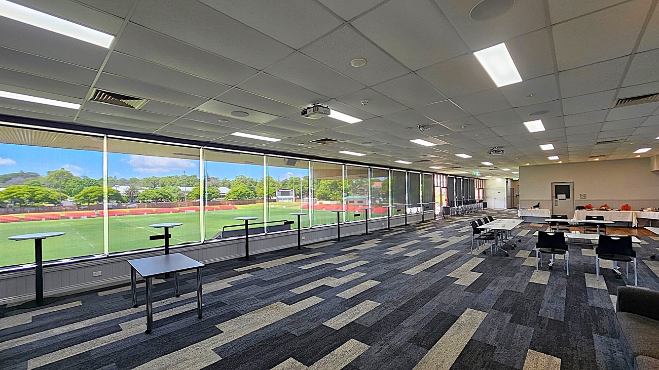 A spacious room with large floor-to-ceiling windows on the left, offering a view of a green sports field and trees outside. The room has a patterned carpet in shades of grey and beige. On the right, there are tables and chairs arranged in rows, with some tables covered in white cloths. The ceiling is fitted with fluorescent lights and a projector.