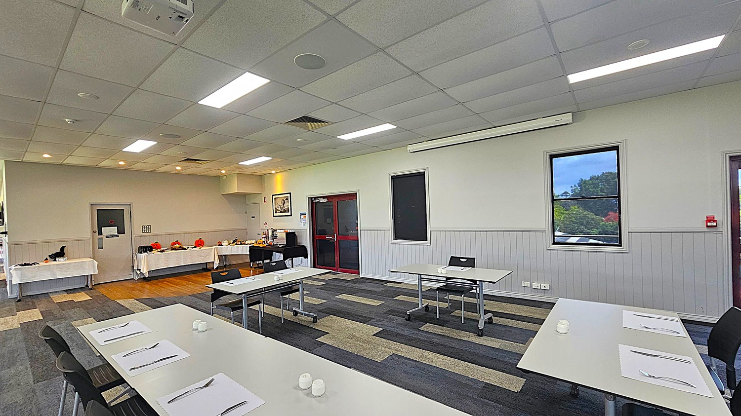 A conference room with a patterned carpet floor and a tiled ceiling with fluorescent lights. On the left, there are tables covered with white cloths, decorated with Halloween-themed items like pumpkins. In the centre, there are several white tables with chairs, each set with placemats and cutlery. The right wall has two windows, one showing a view of trees outside. A red-framed door is also on the right wall. A projector is mounted on the ceiling.