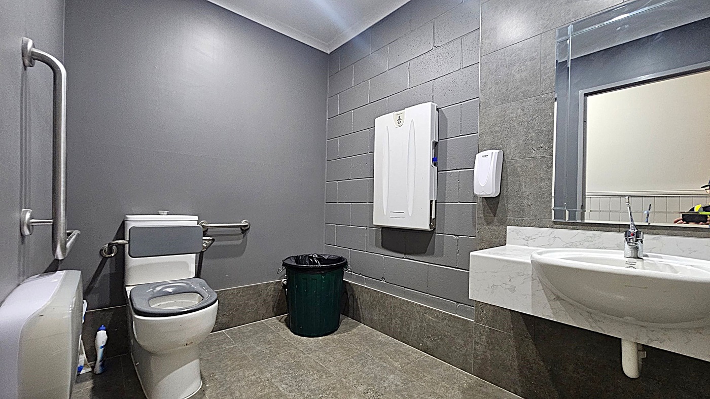 A public restroom with grey walls and a green-tiled floor. On the left, there is a toilet with grab bars and a toilet paper dispenser. In the centre, a black bin is placed against the wall. On the right, a white baby changing station is mounted on the wall above a soap dispenser. A white sink with a chrome tap is below a large mirror. The ceiling is white with recessed lighting.
