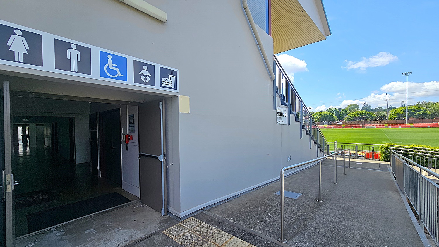 On the left, there is an entrance with signs indicating facilities for women, men, disabled access, baby changing, and food. The entrance is dark inside. On the right, a ramp with metal railings leads down to a sports field with green grass and red seating. A staircase is visible above the ramp. In the background, there are trees and a clear sky.