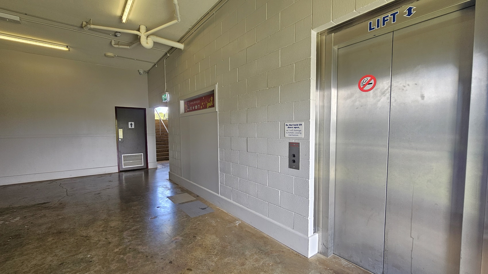 A corridor with a lift on the right, featuring metallic doors and a no-smoking sign. The wall is made of light-coloured bricks. On the left, there's a door with a sign indicating a toilet, and an emergency exit sign above it. In the background, a staircase leads upwards. The floor is a polished concrete, and the ceiling has exposed pipes and fluorescent lights.