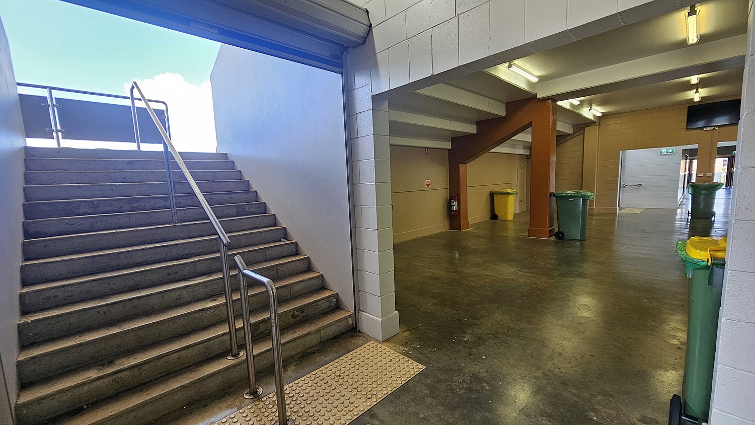 On the left, a concrete staircase with metal railings leads upwards, with a tactile paving strip at the base. On the right, a corridor with a polished concrete floor features several green and yellow rubbish bins. The ceiling has fluorescent lights, and a fire extinguisher is mounted on the wall. An open doorway at the end of the corridor reveals another room.