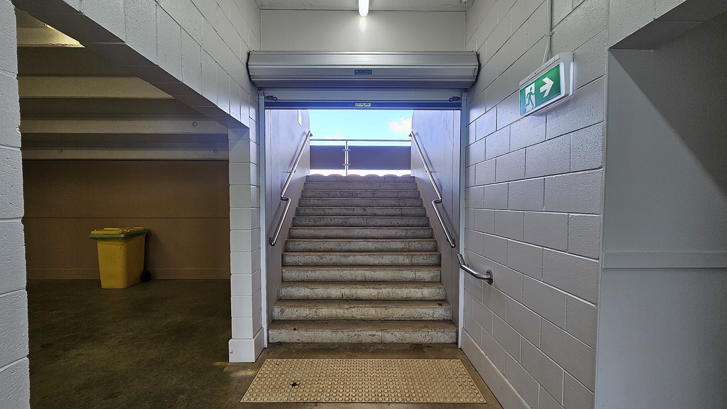 A stairway with metal handrails on both sides leads upwards to an outdoor area. The walls are made of light grey bricks. On the left, there's a yellow bin against the wall. An emergency exit sign with a green background and white arrow is mounted on the right wall. The floor is concrete with a textured mat at the base of the stairs. The ceiling is plain with a fluorescent light fixture.