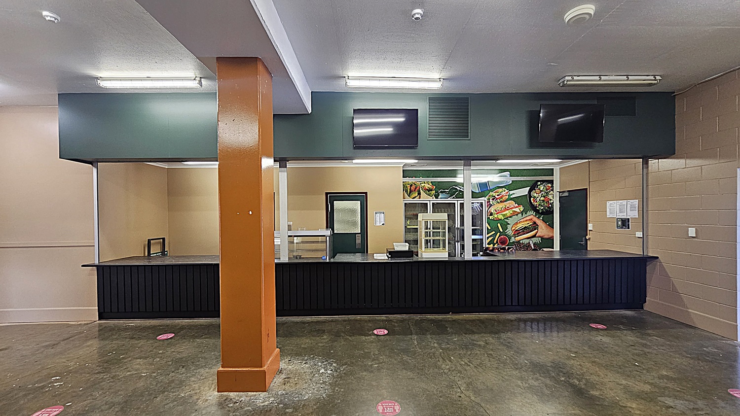 A cafeteria counter with a black front and a green top spans the width of the image. Behind the counter, there's a door and a display case. The wall features a large food poster with images of burgers and salads. An orange column stands in the foreground. The floor is concrete with red social distancing markers, and the ceiling is white with fluorescent lights. Two black screens are mounted above the counter.