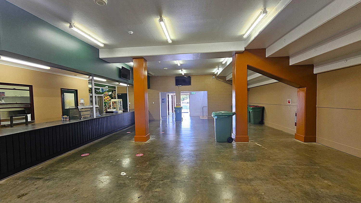 A spacious indoor area with a polished concrete floor. On the left, there's a long counter with a dark front and a service area behind it, featuring a mural of food. The ceiling has fluorescent lights. In the centre, two orange pillars support the ceiling. At the far end, a doorway leads outside, with a green bin nearby. On the right, a sloped ceiling with exposed beams and more bins along the wall.