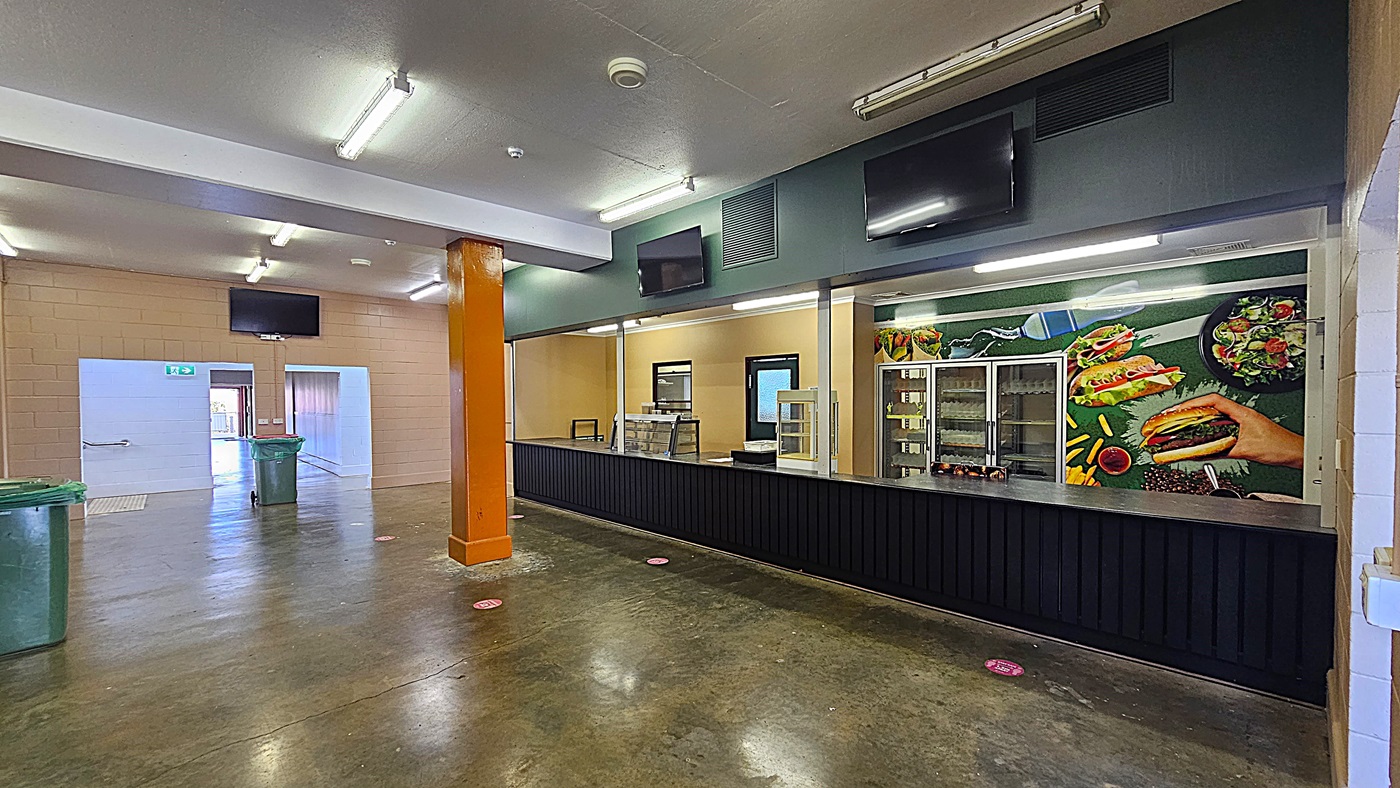 A spacious indoor area with a polished concrete floor and fluorescent lighting on the ceiling. On the right, there's a long black counter with a colourful mural of food items like burgers and salads behind it. Refrigerated display units are visible. On the left, there are two green bins and a doorway leading to another room. An orange pillar stands near the centre, and two black screens are mounted above the counter.