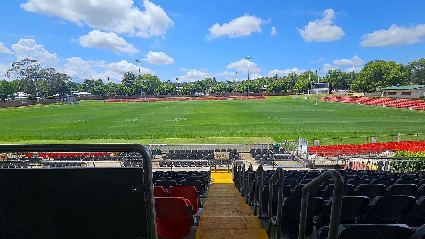 A rugby field with lush green grass is surrounded by red seating on the left and right. In the foreground, there are rows of black and red seats leading down to the field, with a central staircase. The background features trees and a few buildings beyond the field. Two sets of rugby posts are visible at each end of the field.